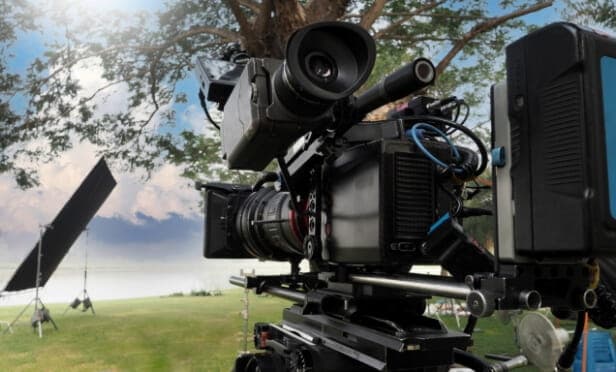 A professional video camera set up outdoors on a grassy area, with lighting equipment and a large tree in the background, captures the essence of brand production under a partly cloudy sky.
