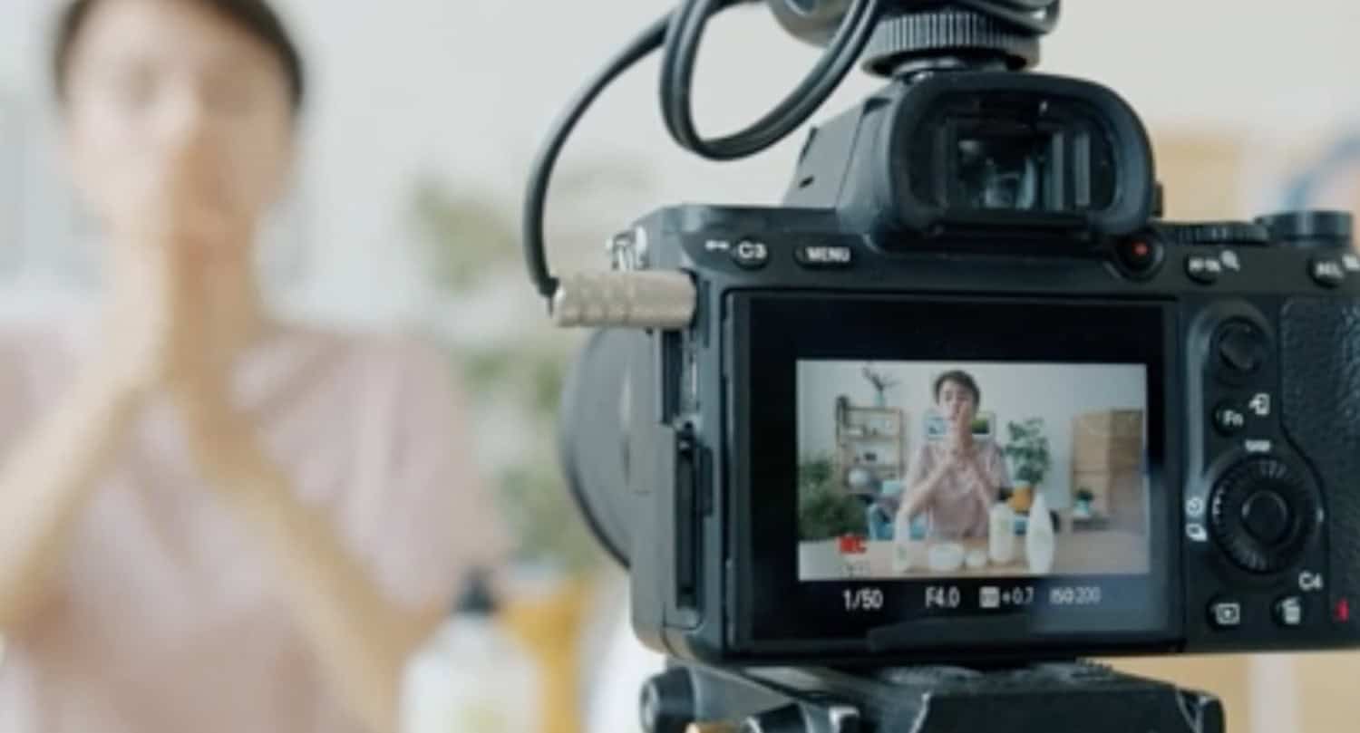 A digital camera on a tripod records a person indoors, likely creating marketing content. The camera screen shows the brand's subject sitting at a table, gesturing with their hands, while appearing slightly blurred in the background.