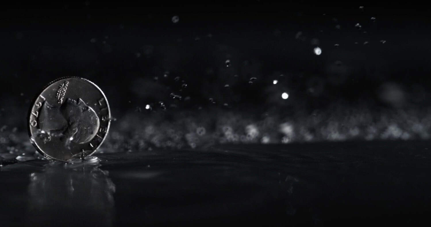 A coin standing upright on a wet surface with water droplets splashing around it, perfect for eye-catching social or marketing video content against a dramatic dark background.