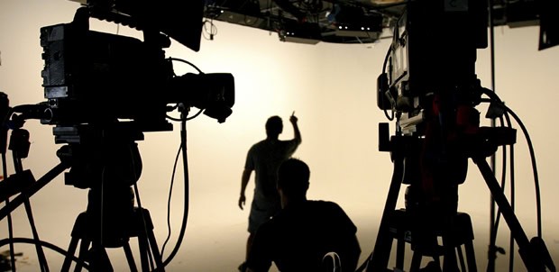 Two video cameras in the foreground film a person standing and gesturing on a brightly lit studio set, capturing a dynamic video production environment. Another person is seated in front of them, their silhouettes highlighting the creative studio atmosphere.