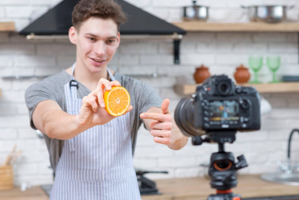 A person wearing an apron holds half an orange and gestures during a video production in a kitchen, with a camera set up on a tripod.
